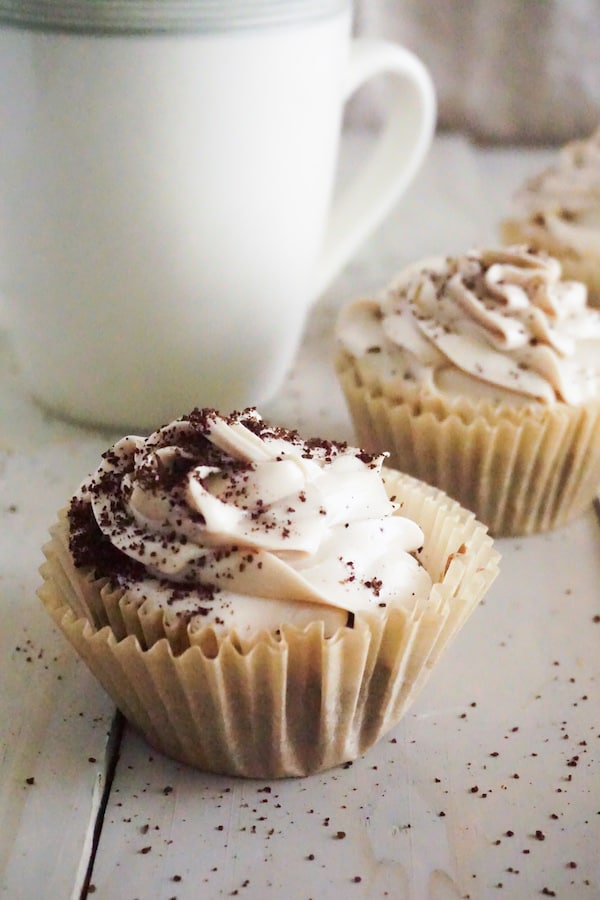 coffee cupcakes topped with frosting and sprinkled with coffee grounds on a table with a cup in the background