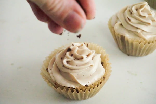 a hand sprinkling coffee grounds on a frosted cupcake