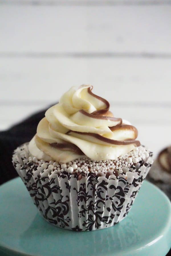 a close up view of nutella cupcake topped with white pearl sprinkles and swirled nutella and white frosting on a green cake stand on a white counter next to a black linen and more cupcakes in the background