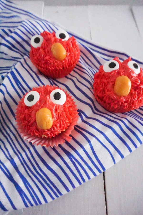 three cupcakes made to look like Elmo with red frosting, candy eyes and an orange starburst as a nose, on a wood table and a white and blue striped linen