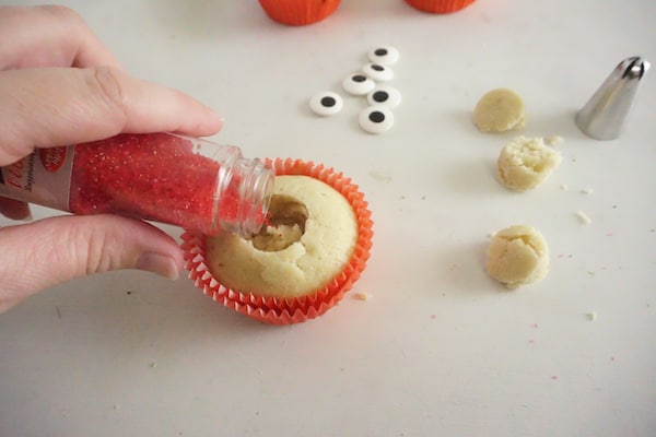 a hand pouring red sanding sugar in the center of a cupcake with candy eyes, cupcake pieces and a decorating tip in the background on a white counter