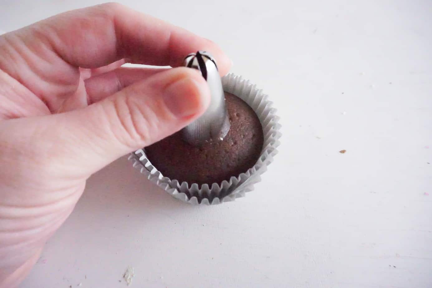 a hand holding a decorating tip over the center of a chocolate cupcake on a white counter