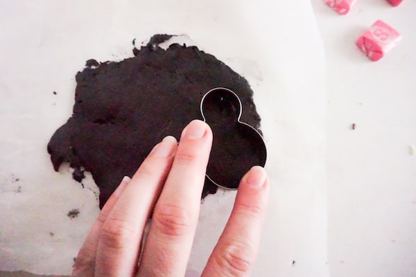 a hand pushing a minnie mouse cookie cutter into black fondant on parchment paper
