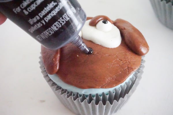 black writing gel being used to draw a mouth on a cupcake decorated with brown and white fondant to look like a dog