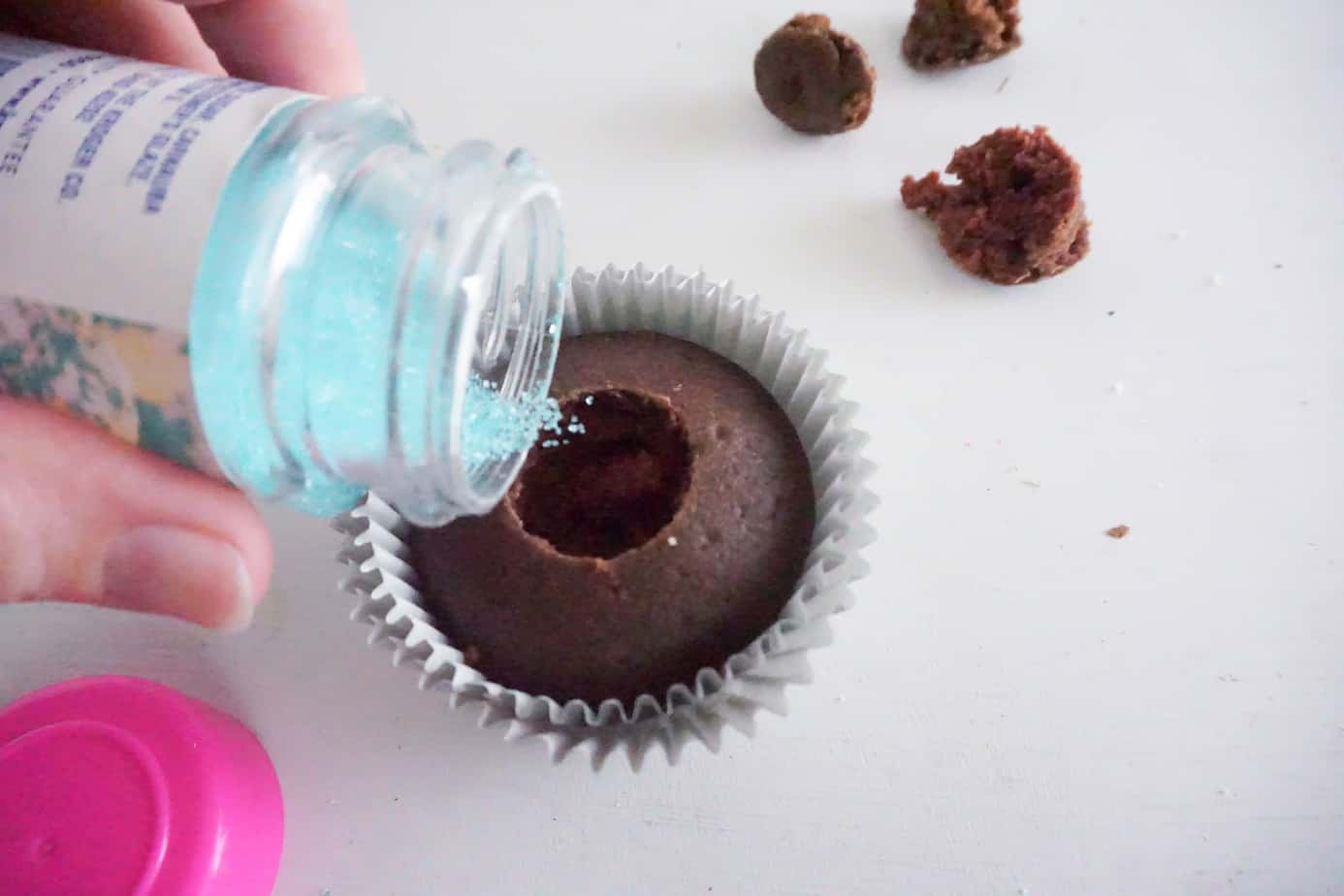 a hand pouring blue sugar sprinkles into a hole in the center of a chocolate cupcake with the rest of the pieces of the cupcake next to it on the white counter 