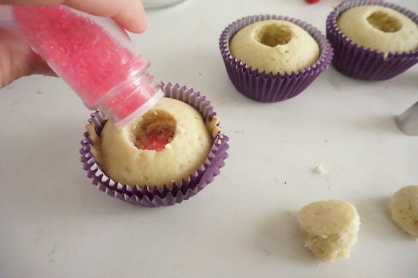 a hand pouring pink sanding sugar into a well of a cupcake with more cupcakes in the background on a white table