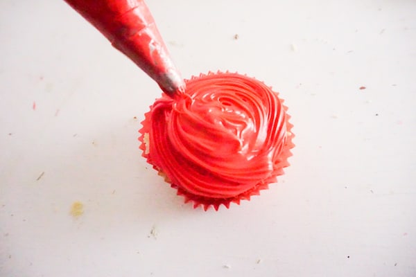 a pastry bag piping red frosting on a cupcake on a white background