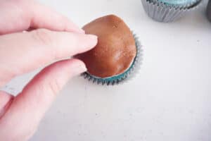 a hand placing brown fondant on a cupcake with another cupcake in the background on a white counter