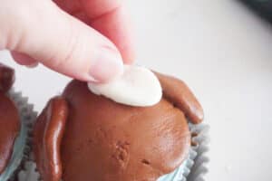 a hand placing a piece of white fondant on a cupcake topped with brown fondant