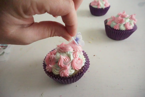 a hand placing an edible butterfly on a cupcake decorated with pink and green icing with more cupcakes in the background on a white table