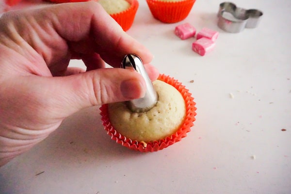 a hand pushing a piping tip into the center of a cupcake to create a well with other cupcakes, pink starbursts and a minnie mouse cookie cutter in the background
