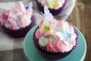 a cupcake with pink and green frosting with edible butterflies on top, on a green cake stand on a brown table with two more cupcakes in the background on a linen