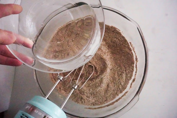 a hand pouring a glass bowl of water into a bowl of cupcake ingredients with a mixer in it