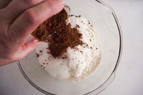 a hand pouring cocoa powder into a glass bowl filled with flour and other dry ingredients