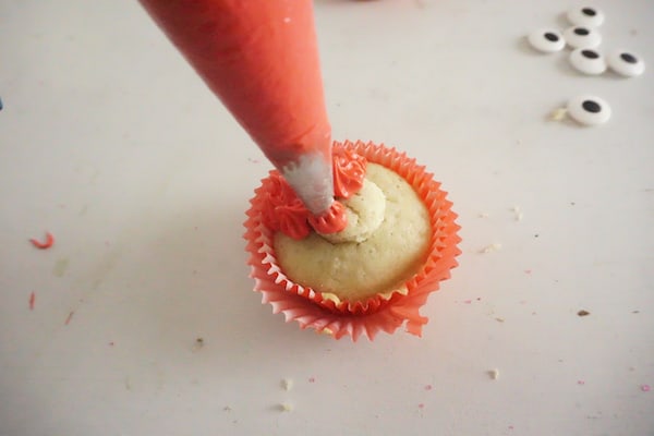 a pastry bag piping red frosting onto a cupcake on a white counter with candy eyes in the background