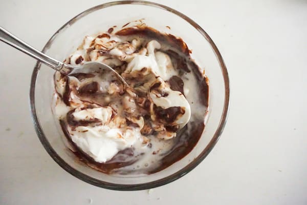coffee grounds, cream, and melted chocolate being mixed by a spoon in a glass bowl on a white table