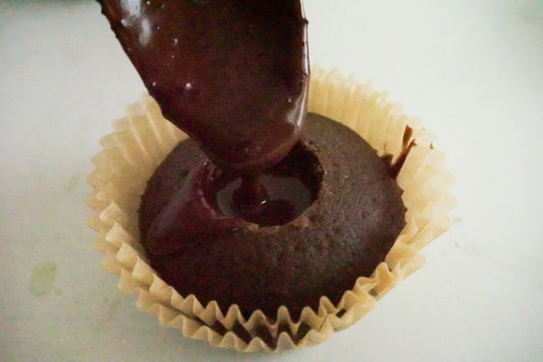 a spoon pouring chocolate ganache in the center of a cupcake