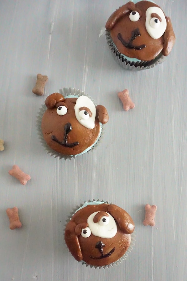 cupcakes topped with brown, white and block frosting to look like the face of a dog next to dog bone treats on a table