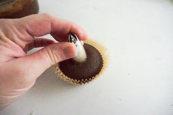 a hand placing a decorating tip in the center of a cupcake on a white table