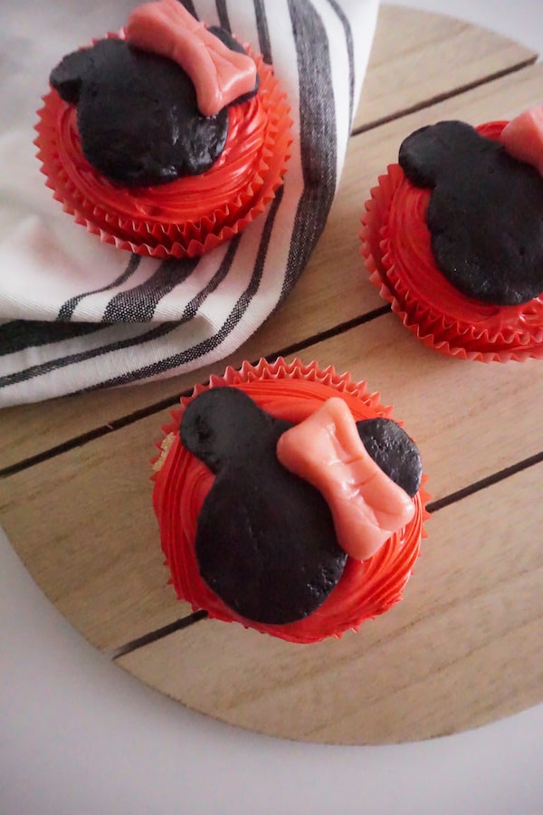 three cupcakes decorated with red frosting, black fondant to look like Minnie Mouse and a pink Starburst shaped to look like a bow, on a wooden trivet on a gray and white cloth, on a white background