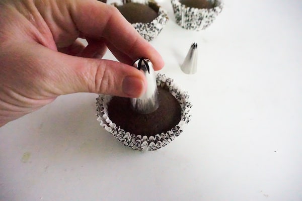 a hand pressing a decorating tip into the top of the cupcake off-center, creating a well with more cupcakes in the background on a white counter