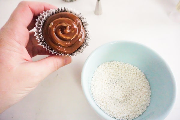 a hand holding a cupcake topped with nutella next to a bowl of white pearl sprinkles on a white counter