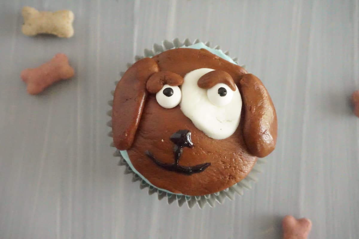 cupcake topped with brown, white and block frosting to look like the face of a dog next to dog bone treats on a table