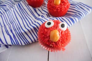 a closeup of a cupcake made to look like Elmo with red frosting, candy eyes and an orange starburst as a nose, on a wood table and a white and blue striped linen with two more cupcakes in the background
