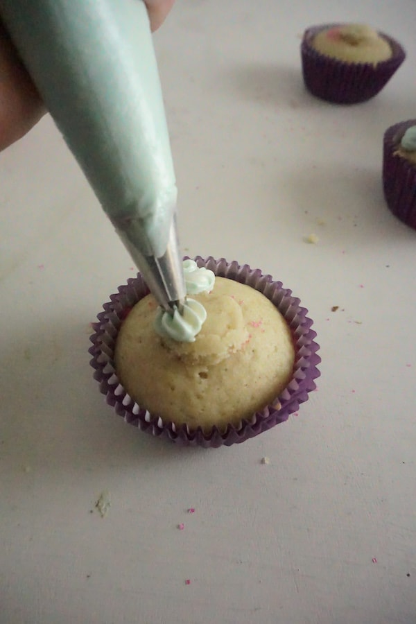 using a pastry bag to pipe frosting on a cupcake with more cupcakes in the background on a white table