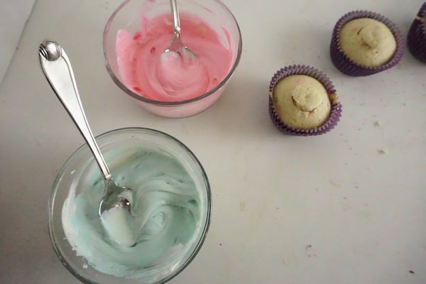 pink and green frosting in glass bowls next to two cupcakes on a white table