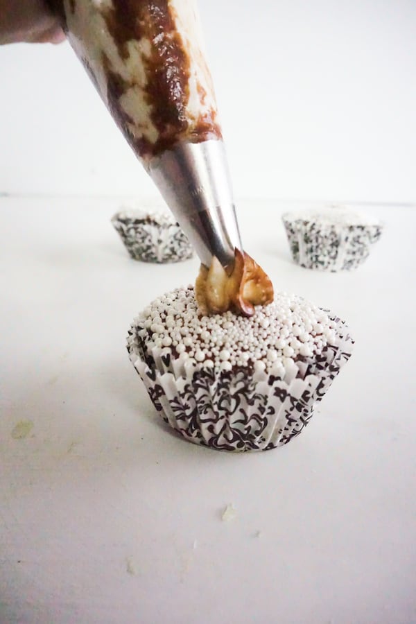 a mix of white frosting and nutella in a pastry bag being piped on a cupcake topped with white pearl sprinkles with more cupcakes in the background on a white counter