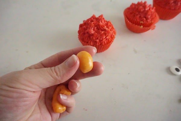 a hand holding orange starburst rolled into the shape of a ball with cupcakes with red frosting in the background on a white counter