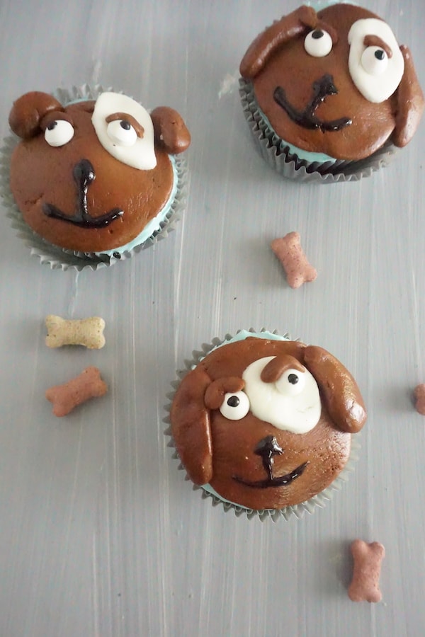 cupcakes topped with brown, white and block frosting to look like the face of a dog next to dog bone treats on a table