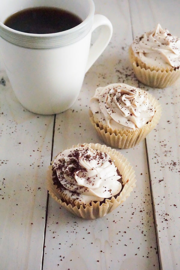 coffee cupcakes topped with frosting and sprinkled with coffee grounds on a table with a cup of coffee next to them
