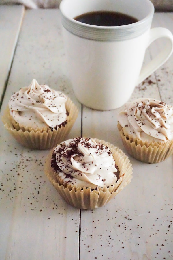 coffee cupcakes topped with frosting and sprinkled with coffee grounds on a table with a cup of coffee in the background