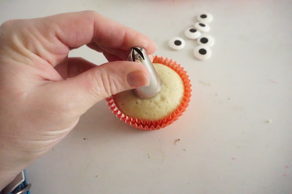 a hand pressing a decorating tip in the center of a cupcake on a white counter with candy eyes in the background