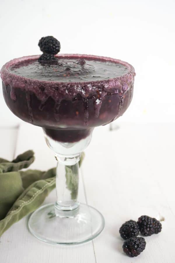side view of a blackberry margarita in margarita glass on a white background with a moss green linen and random blackberries nearby