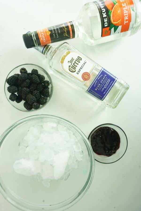 ingredients for making a blackberry margarita (ice, blackberries, blackberry jam, tequila and triple sec) on a white background