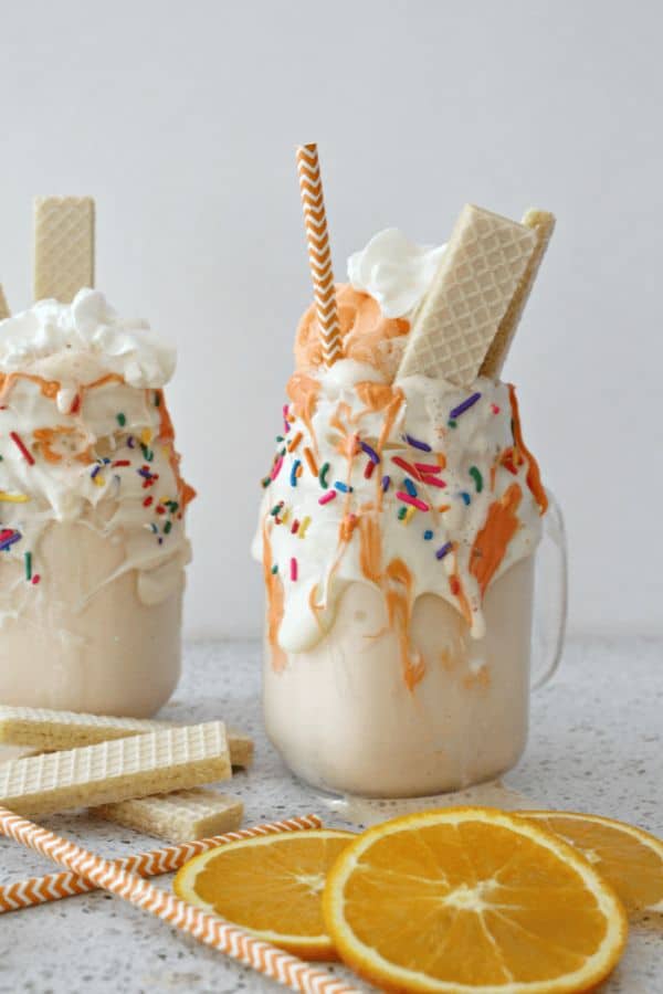 Orange Creamsicle Milkshake in two glasses topped with whipped cream and vanilla wafer cookies with more cookies, orange slices and straws on the gray counter