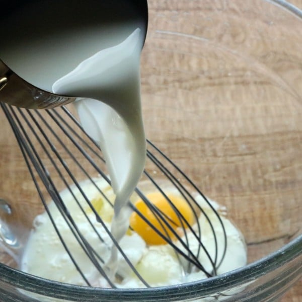 milk being poured from a metal bowl into a glass bowl with eggs and a wire whisk in it