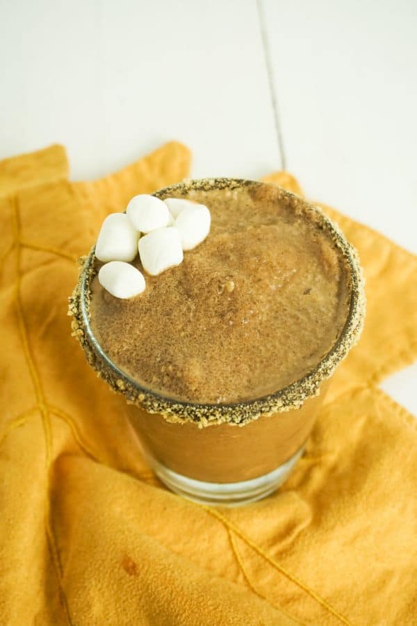 top view of s'mores margarita in glass on orange dish towel, glass rimmed with chocolate and graham cracker crumbs, drink garnished with mini marshmallows