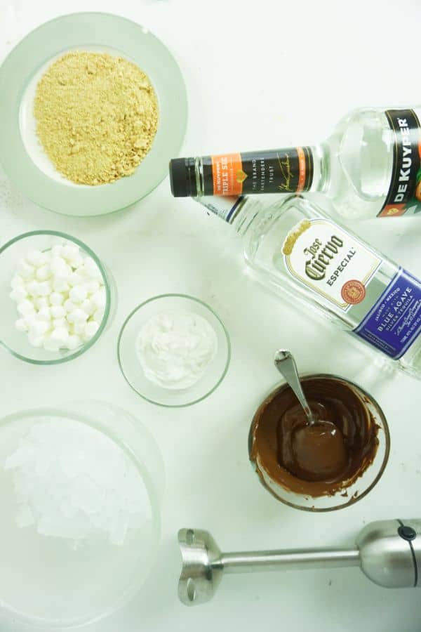 bottles of tequila and triple sec, bowls of coconut cream, melted chocolate chips, ice, crushed graham crackers, marshmallows, immersion blender all on a white table