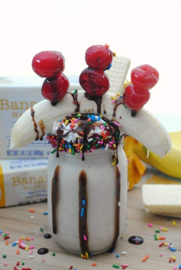 banana split freakshake in a glass topped with a banana, cherries and banana wafer cookies with more cookies and sprinkles in the background on a wood table