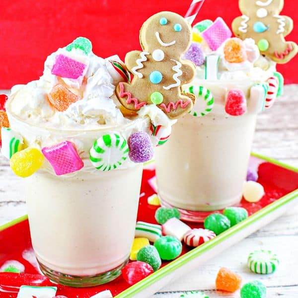 gingerbread house milkshake in two glasses topped with a gingerbread man and colorful hard candies on a red tray with more candies on the tray