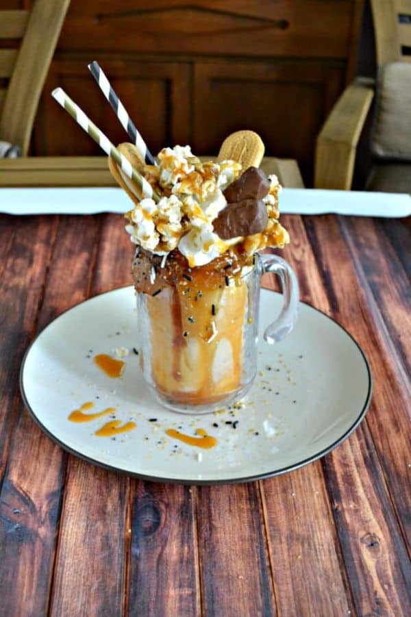 sea salt caramel cookie monster milk shake in a glass topped with homemade caramel corn, nutter butters, Twix, whipped cream, and loads of caramel on a white plate on a wood table