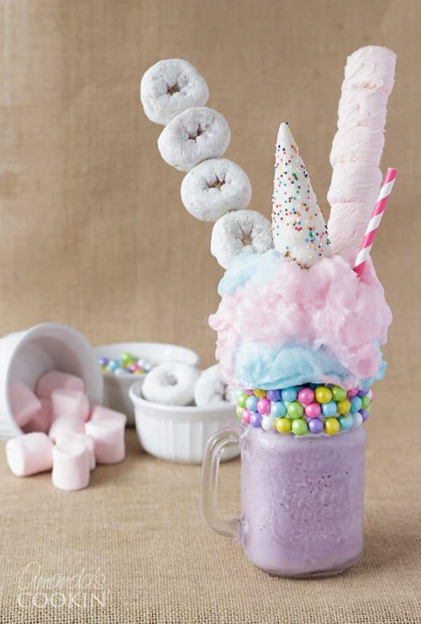 unicorn freakshake by Amandas Cookin in a glass topped with donuts and cotton candy with more donuts and marshmallows in the brown background