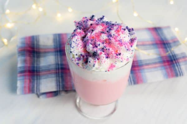 pink hot chocolate topped with whipped cream and colored sprinkles to look like sugar plum hot chocolate with a cloth and lights in the background