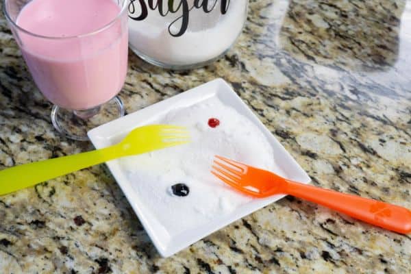 a glass of strawberry milk, a jar of sugar, a plate topped with sugar and drops of food coloring and two forks all on a kitchen counter