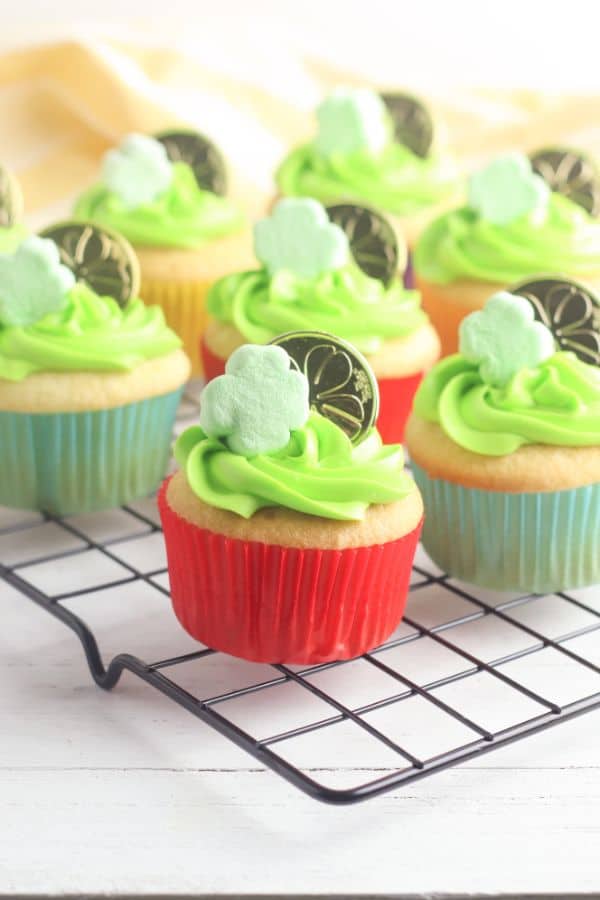 white cupcakes decorated with green frosting, a shamrock marshmallow and gold coin, on a wire rack