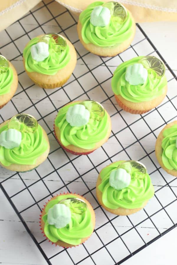 white cupcakes decorated with green frosting, a shamrock marshmallow and gold coin,on a wire rack